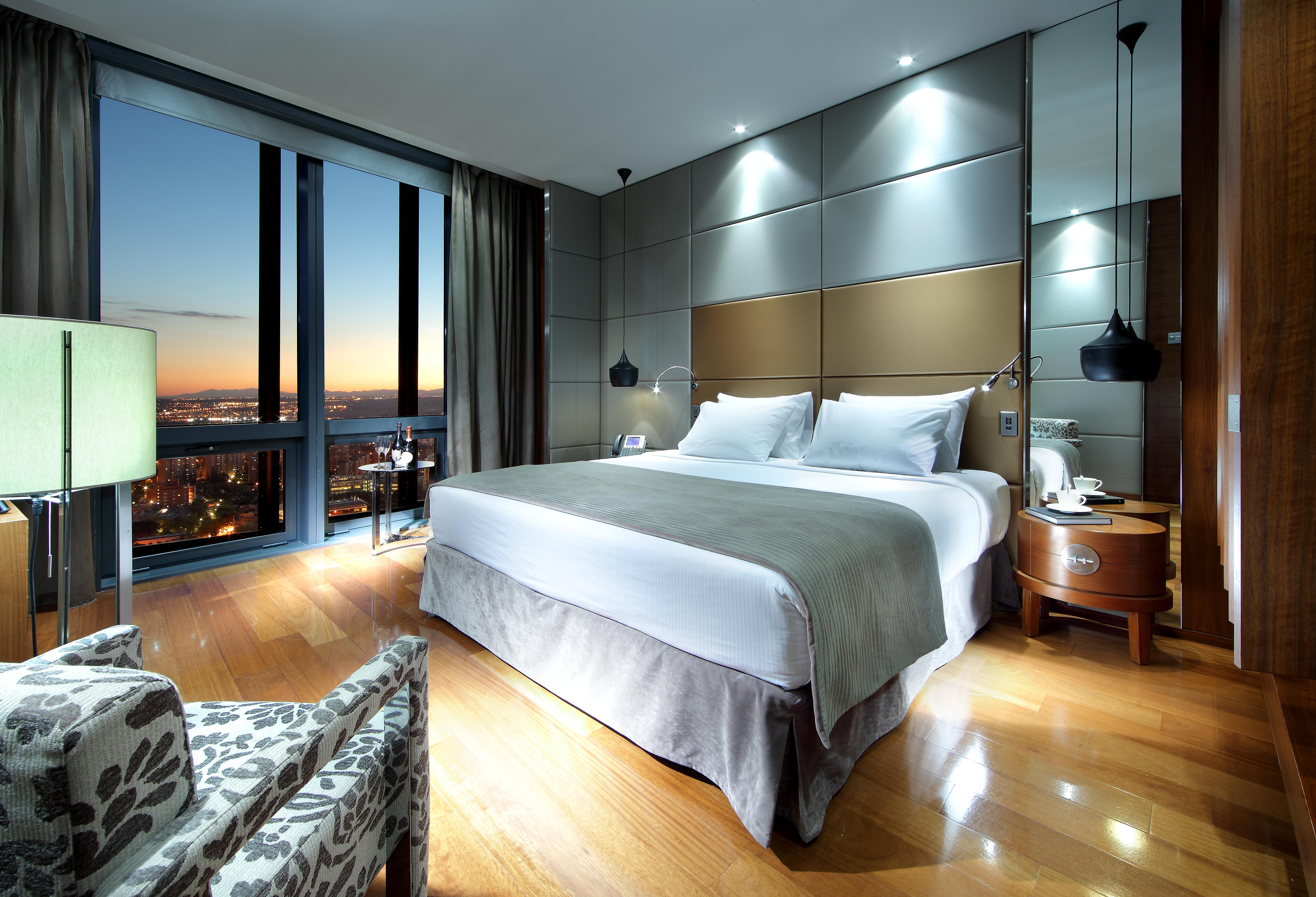Eurostars Madrid Tower Hotel Exterior photo The image shows a modern hotel room featuring a large bed with white linens and a gray throw. There are stylish bedside lamps and a sleek nightstand. A comfortable chair with a patterned fabric sits to the side. The room has large windows that reveal