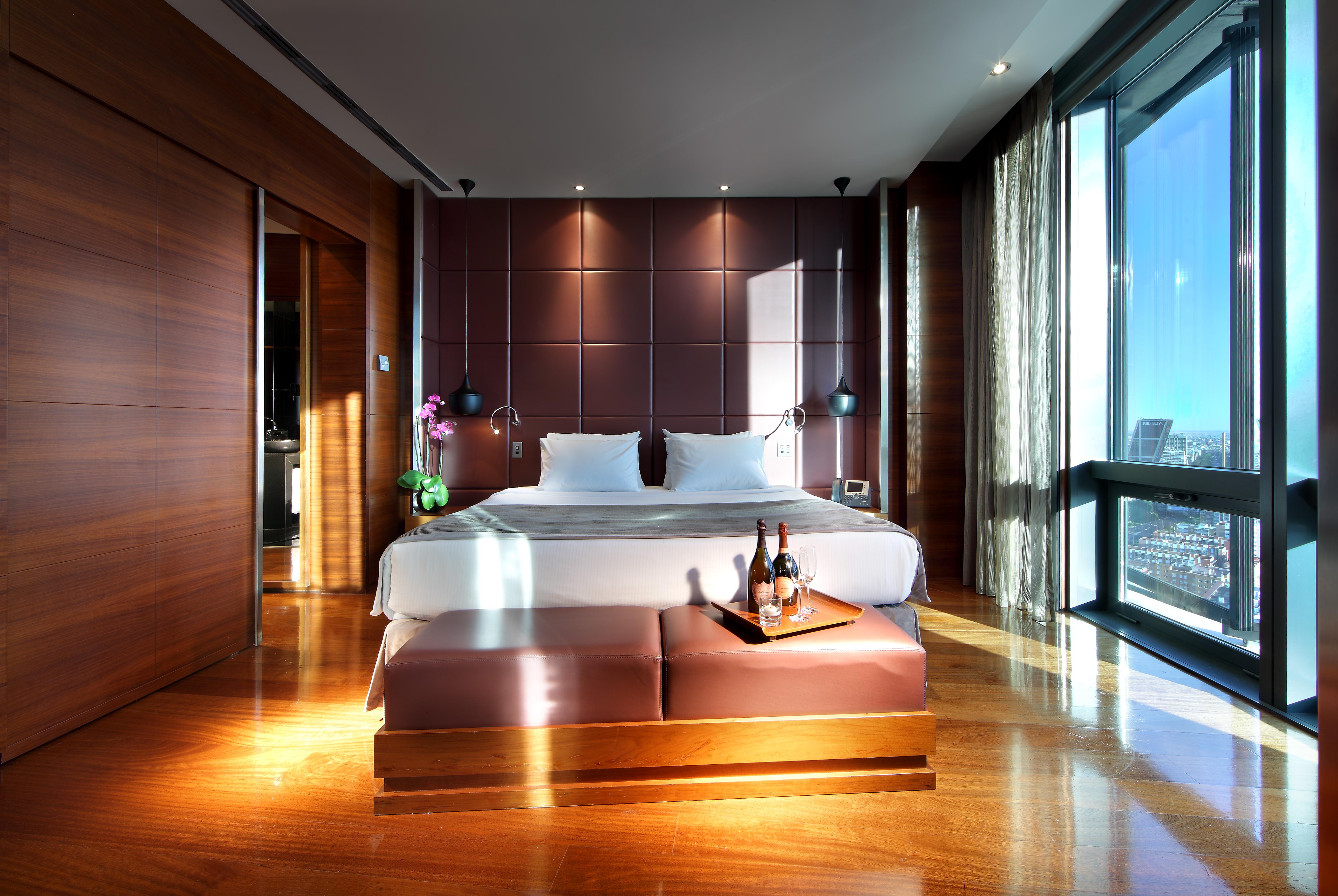 Eurostars Madrid Tower Hotel Exterior photo The photo shows a modern hotel room featuring a large bed with white linens positioned against a stylish dark brown accent wall. There are two bedside lamps on either side of the bed, and a small table at the foot of the bed holding a bottle of wine 