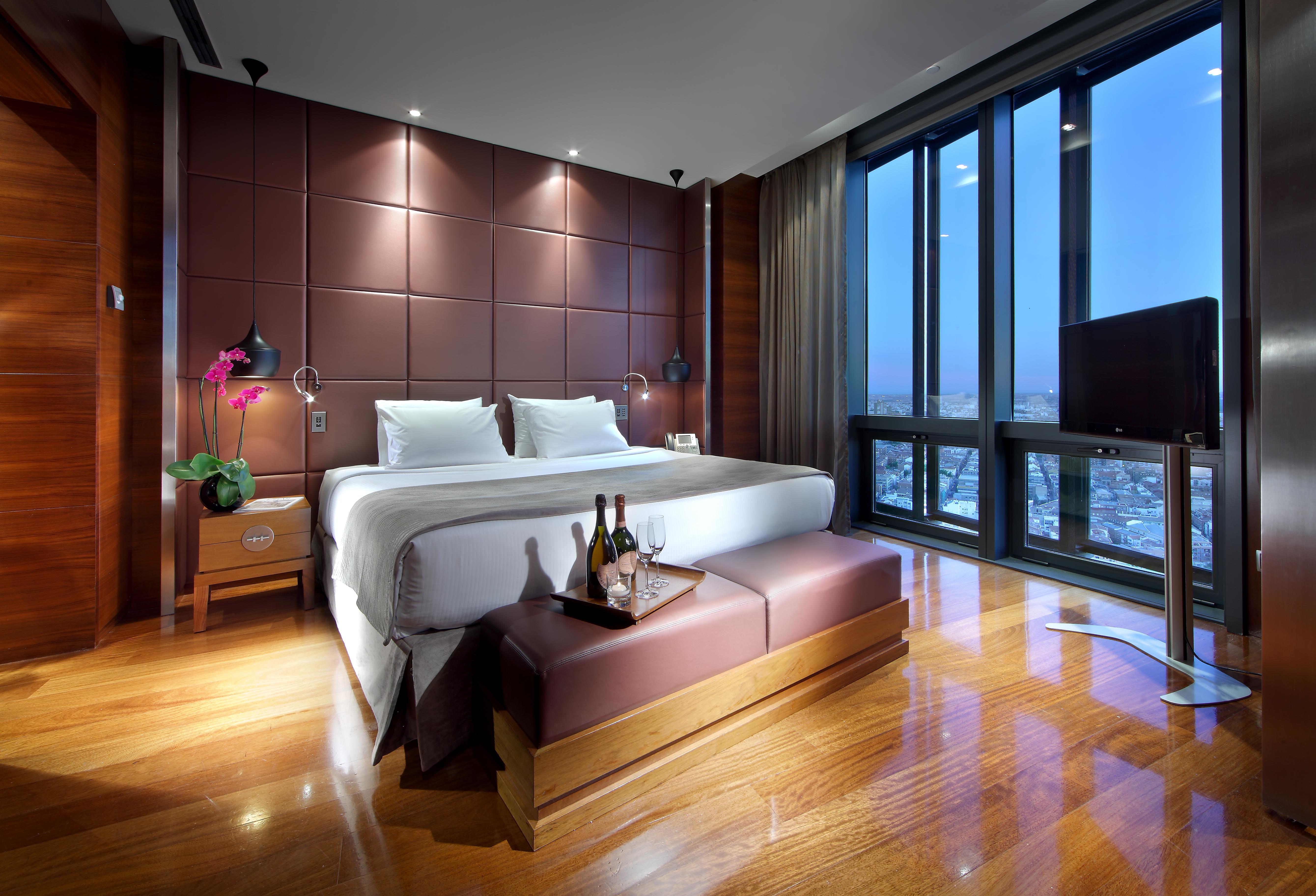 Eurostars Madrid Tower Hotel Exterior photo The photo shows a modern hotel room with a luxurious design. It features a large bed with white linens and plush pillows, positioned against a stylish brown accent wall. There are bedside tables on each side of the bed, each equipped with a lamp. In 