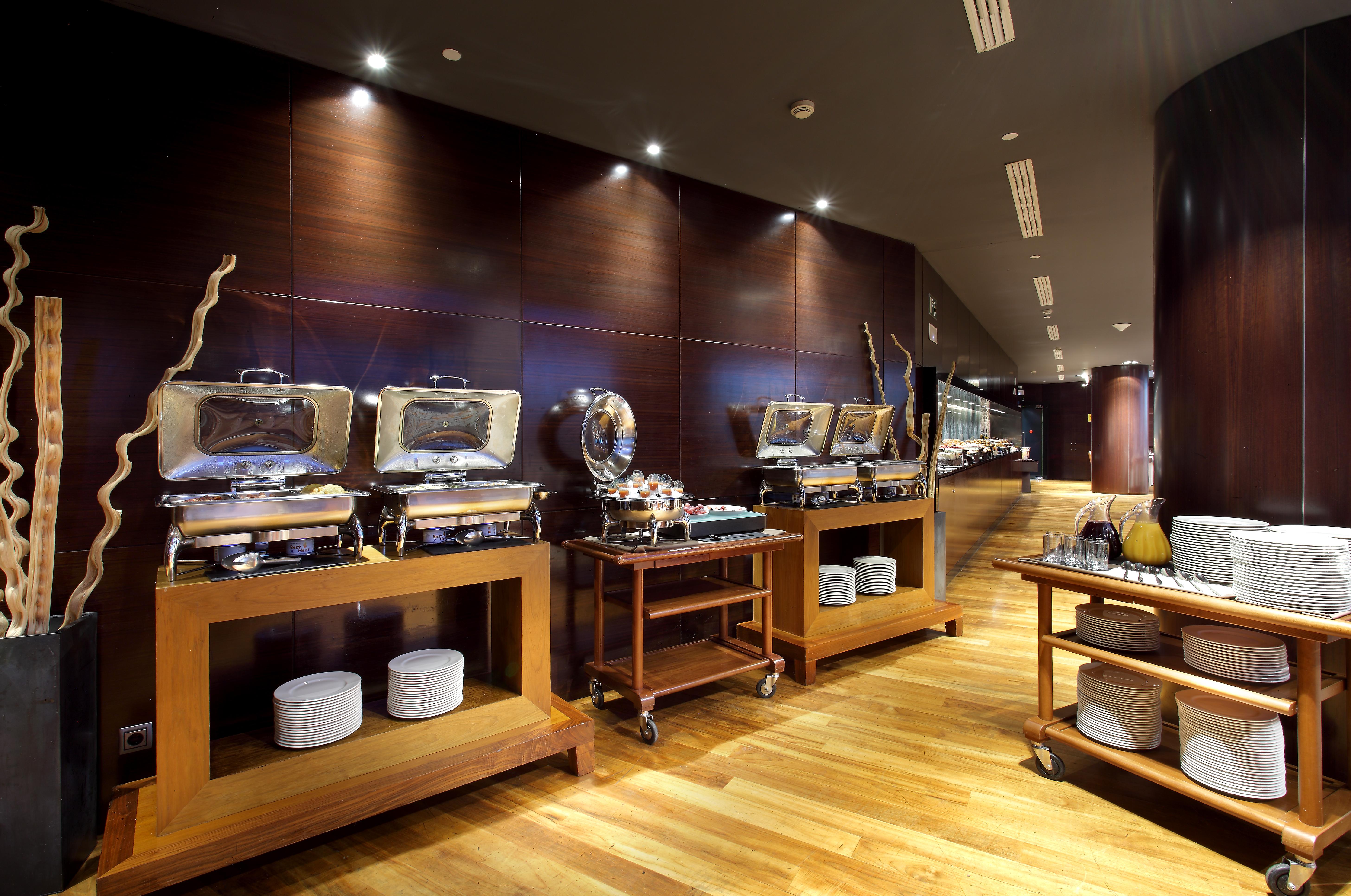 Eurostars Madrid Tower Hotel Exterior photo The photo shows a modern restaurant buffet setting. There are several food warmers placed on wooden tables, showcasing a variety of dishes. The walls have a dark wood finish, and the lighting is soft and ambient, creating an elegant atmosphere. In fr