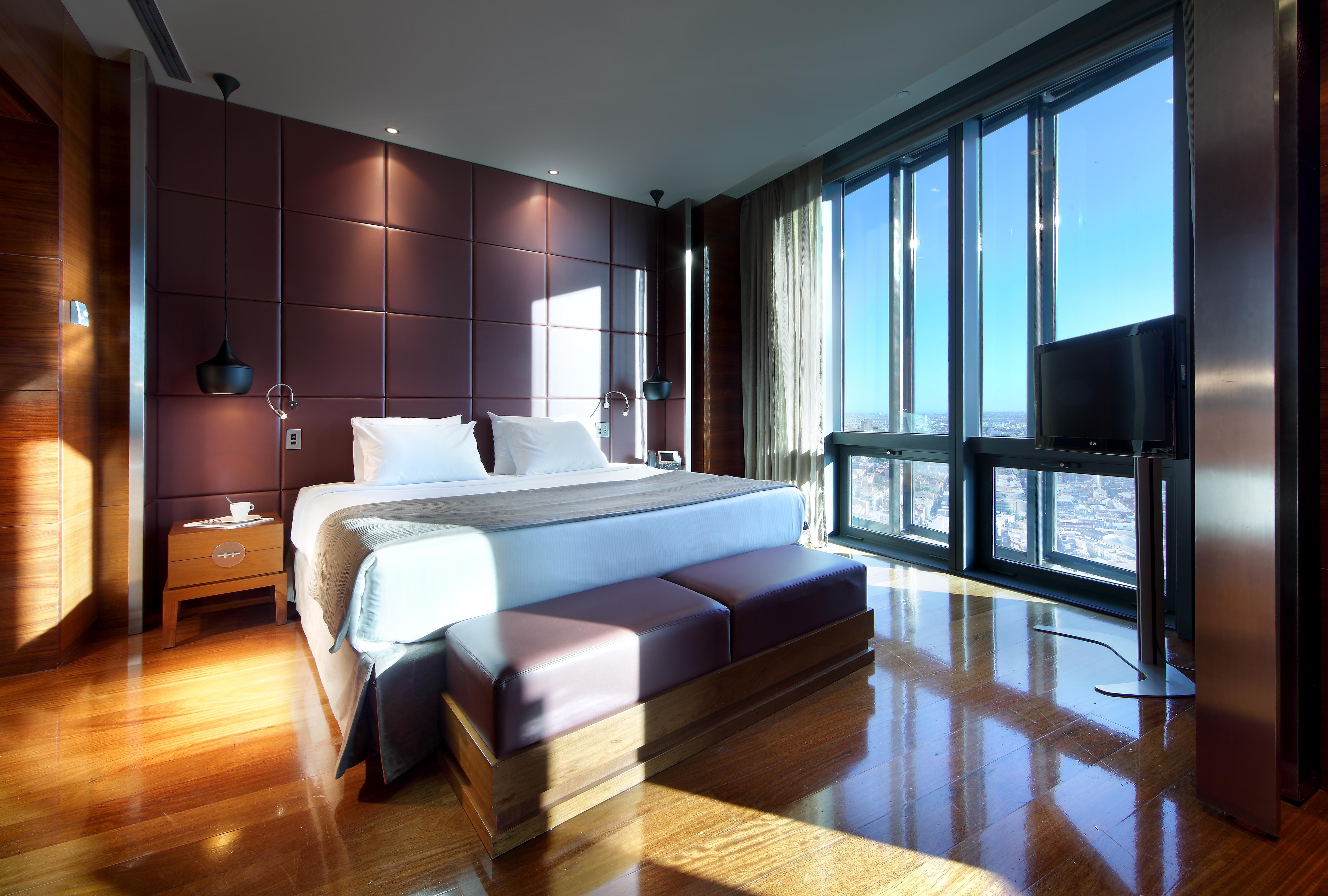 Eurostars Madrid Tower Hotel Exterior photo The photo shows a modern hotel room with sleek design features. It includes a large bed with white bedding, positioned against a wall with dark, textured panels. On either side of the bed, there are bedside tables with lamps. A stylish bench sits at 