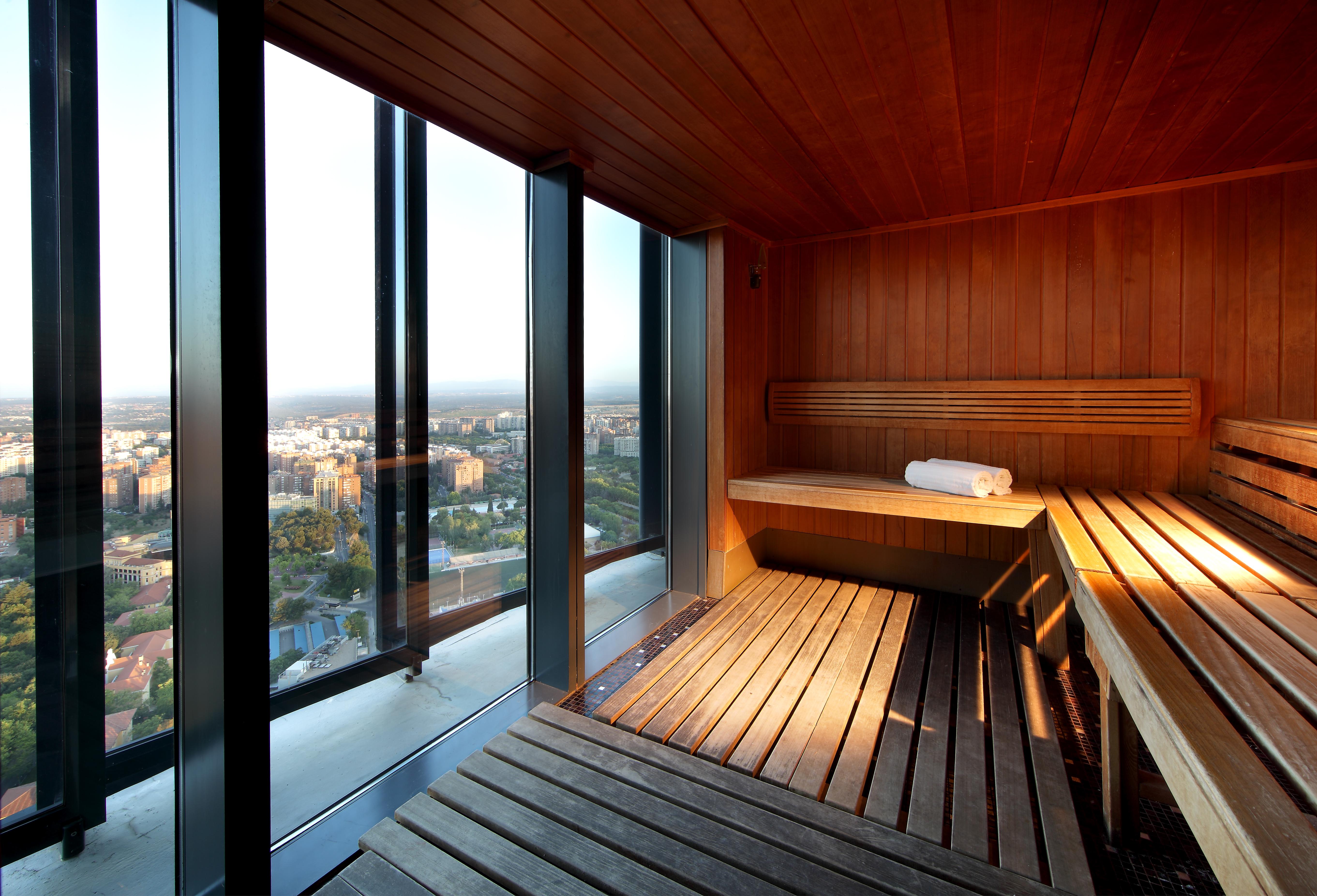 Eurostars Madrid Tower Hotel Exterior photo The image shows a modern sauna with wooden interiors. The sauna features benches made of wood, providing a relaxing space. One of the benches has a rolled towel resting on it. Large glass windows allow for a panoramic view of the outside, showcasing 