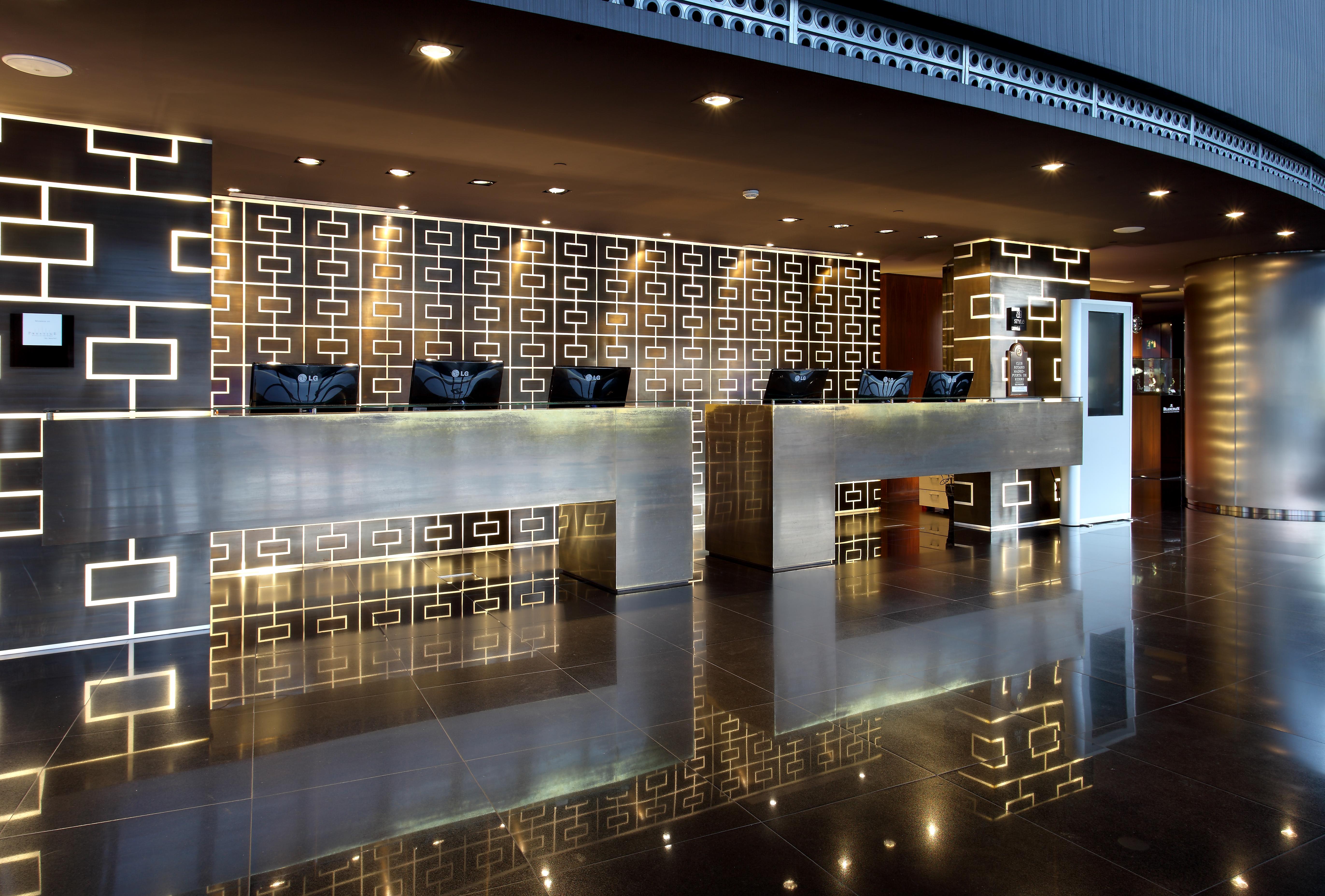 Eurostars Madrid Tower Hotel Exterior photo The photo shows a modern hotel reception area. There are two prominent reception counters made of a sleek, shiny material. The background features a decorative wall with a geometric pattern, possibly in darker colors. The lighting is soft and stylish