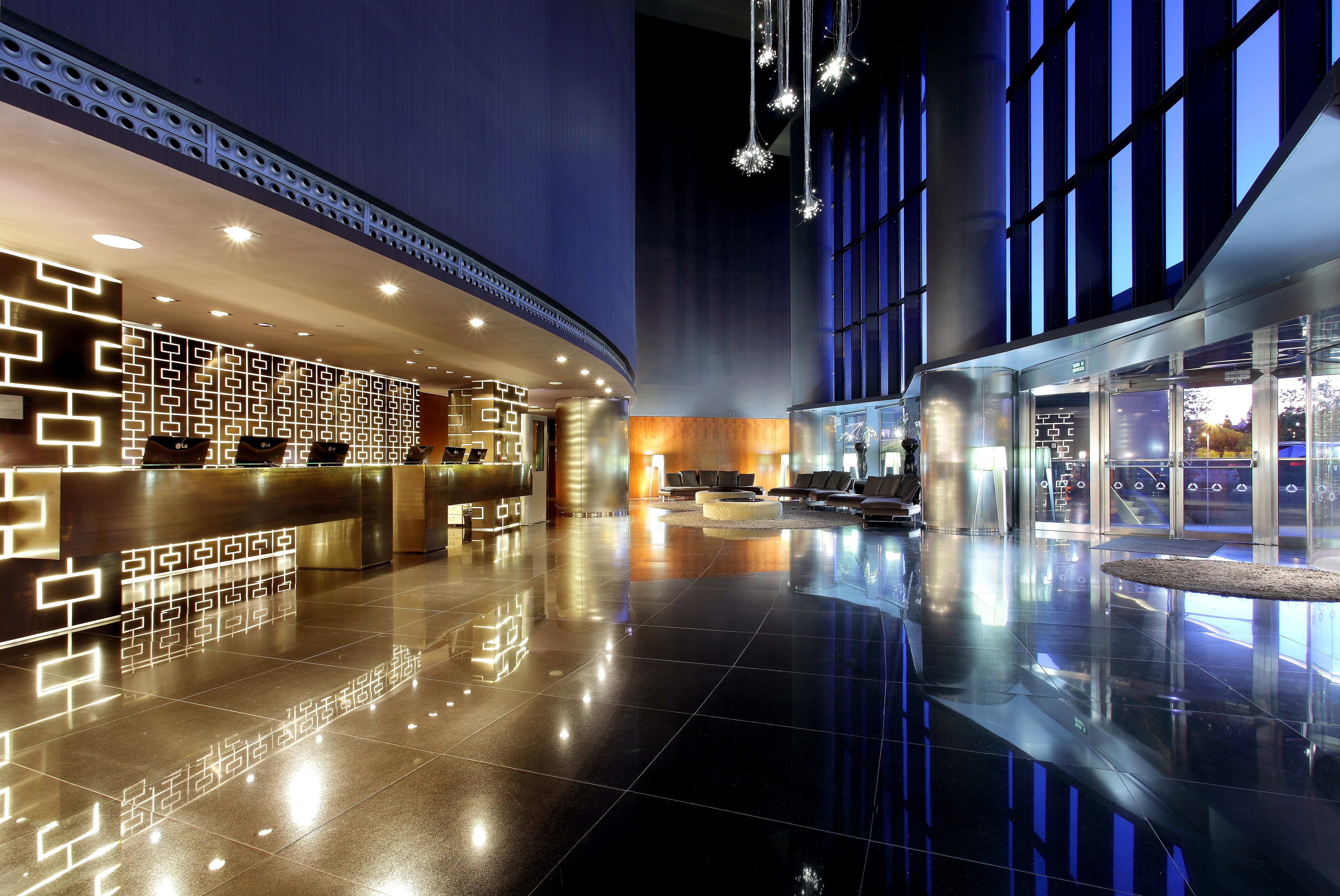 Eurostars Madrid Tower Hotel Exterior photo The image depicts a modern hotel lobby featuring sleek, contemporary design elements. On one side, there is a reception desk with a striking geometric pattern, illuminated with soft lighting. The flooring is glossy and reflective, adding to the elega