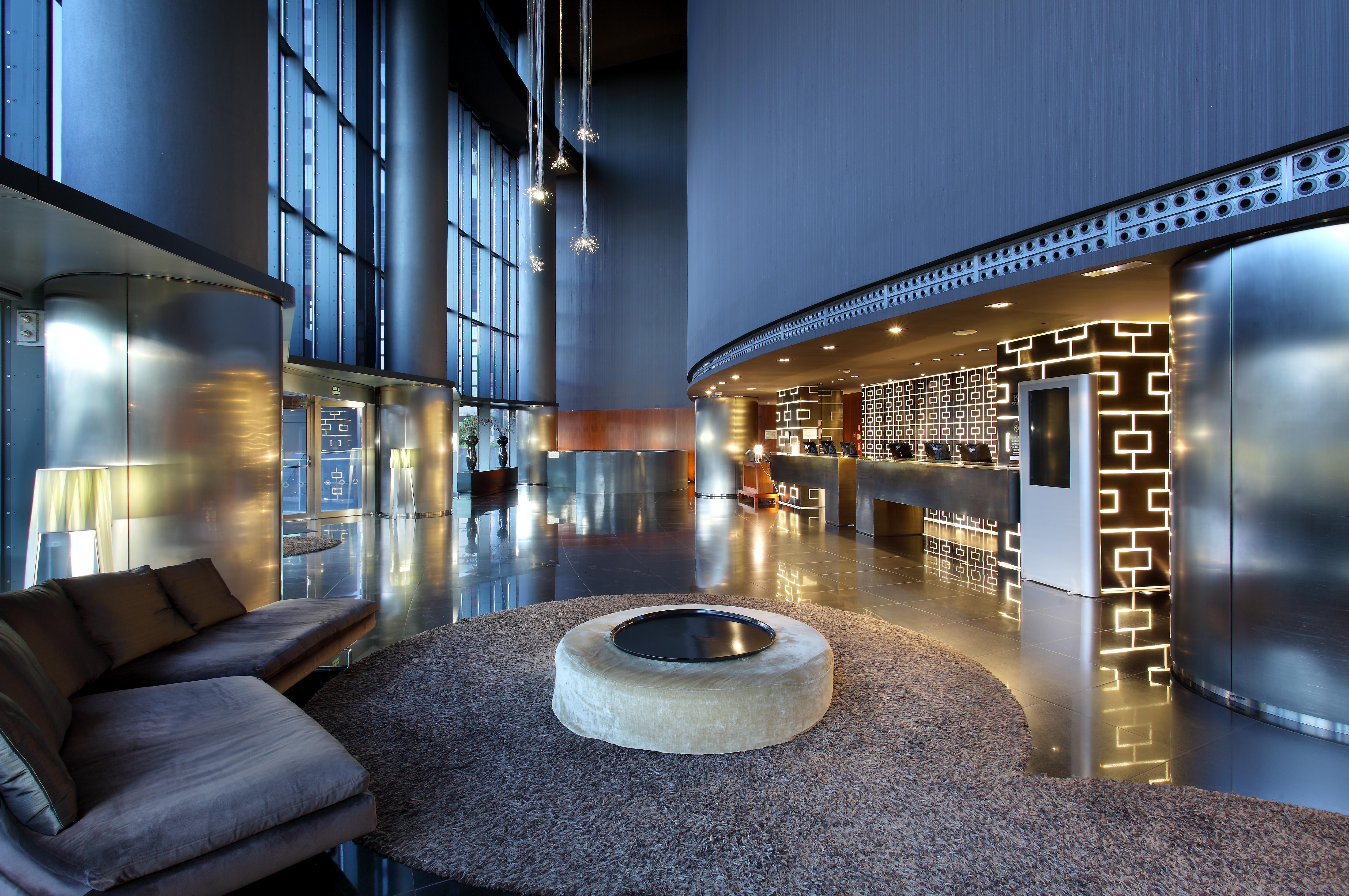 Eurostars Madrid Tower Hotel Exterior photo The photo shows a modern hotel lobby with a sleek and sophisticated design. There is a large, open space featuring tall glass windows allowing natural light to flood in. The reception desk is stylishly designed, with a decorative wall behind it that 