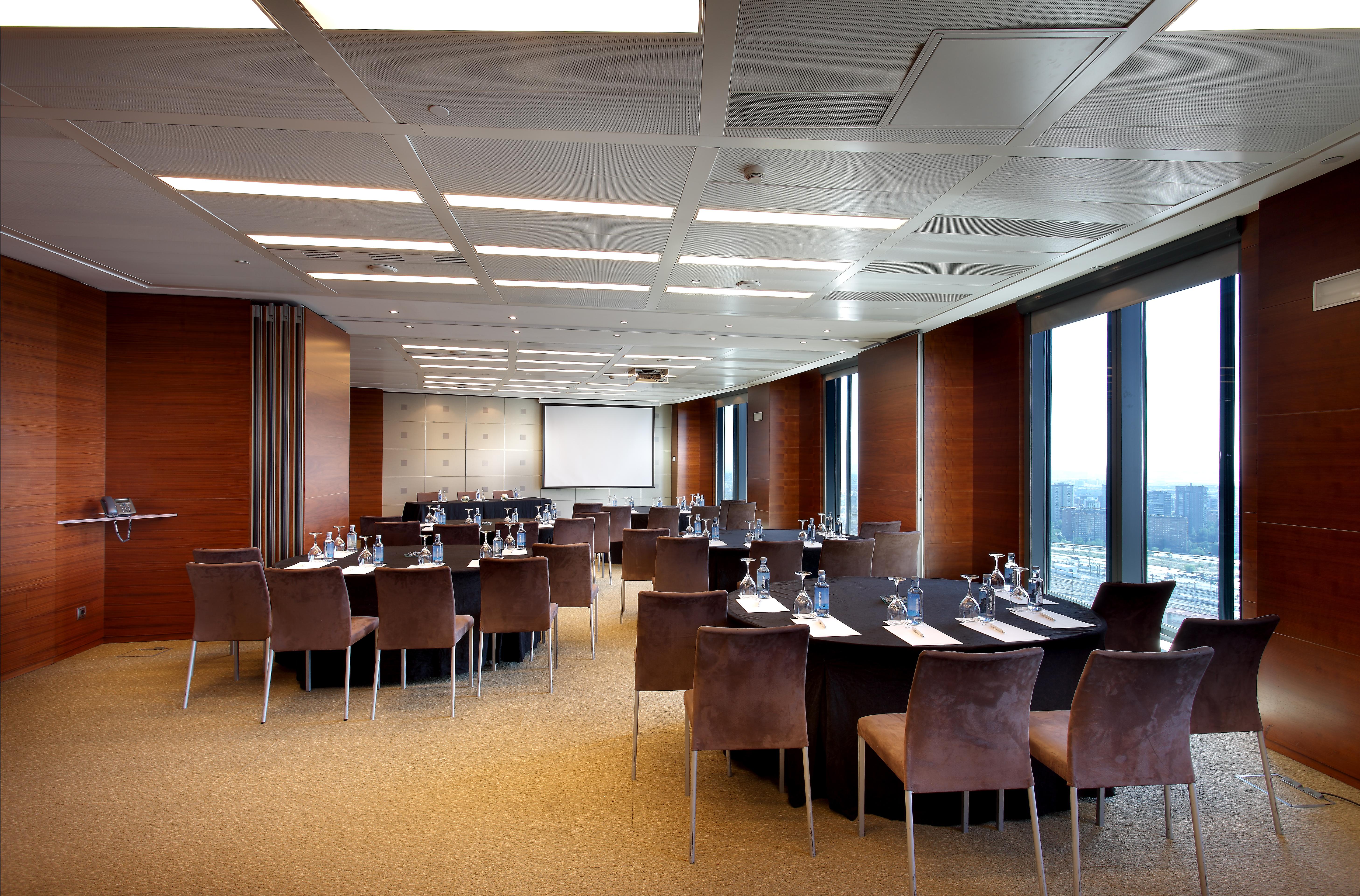 Eurostars Madrid Tower Hotel Exterior photo The photo shows a modern conference room set up for a meeting or event. The room features a large, rectangular table surrounded by upholstered chairs. Each place at the table has a notepad and pen, and there are glasses of water provided. Large windo