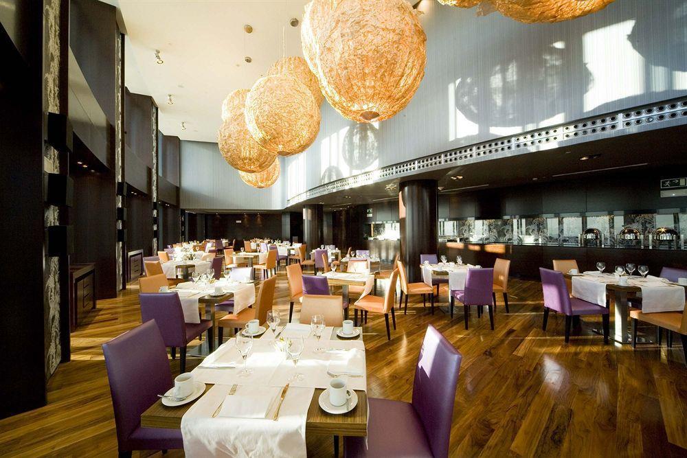 Eurostars Madrid Tower Hotel Restaurant photo The photo shows a modern restaurant interior featuring elegant decor. The dining area includes tables set for guests with white tablecloths, plates, and cutlery. The seating consists of a mix of wooden chairs, some upholstered in purple fabric. The l