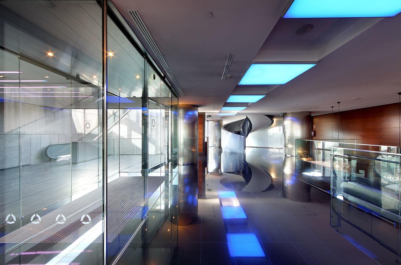 Eurostars Madrid Tower Hotel Exterior photo The photo shows a modern, sleek interior space characterized by large glass walls and reflective surfaces. The ceiling features blue-lit sections, creating a contemporary atmosphere. The polished floor enhances the overall brightness of the area, and
