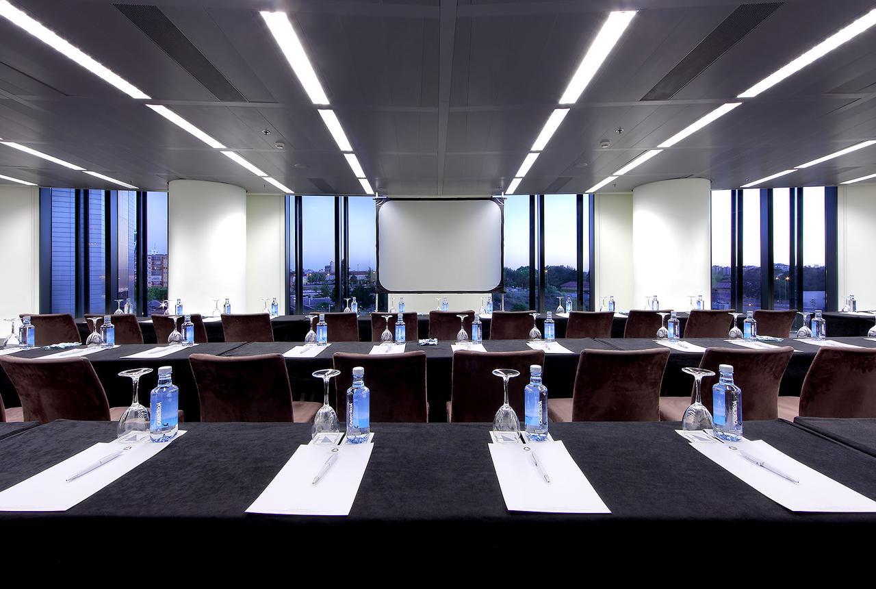 Eurostars Madrid Tower Hotel Exterior photo The photo shows a modern conference room with large windows that provide a view of the cityscape outside. The room is arranged with several tables set up in a classroom or boardroom style. Each table has a black tablecloth and features a glass of wat