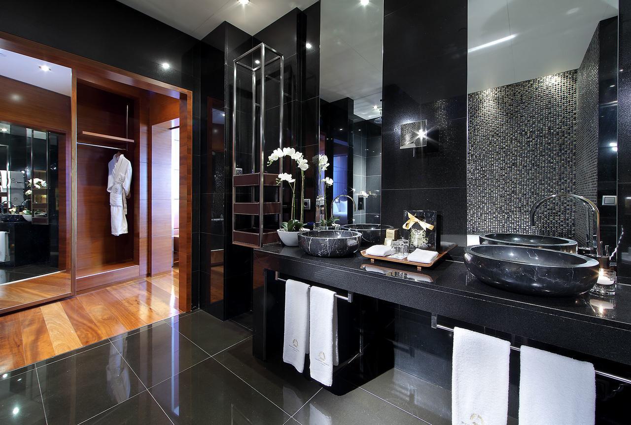 Eurostars Madrid Tower Hotel Exterior photo The photo shows a modern and elegant bathroom with a contemporary design. It features dual black stone sinks with sleek faucets positioned on a dark countertop. The walls are adorned with shiny black tiles and a mosaic pattern, contributing to a luxu