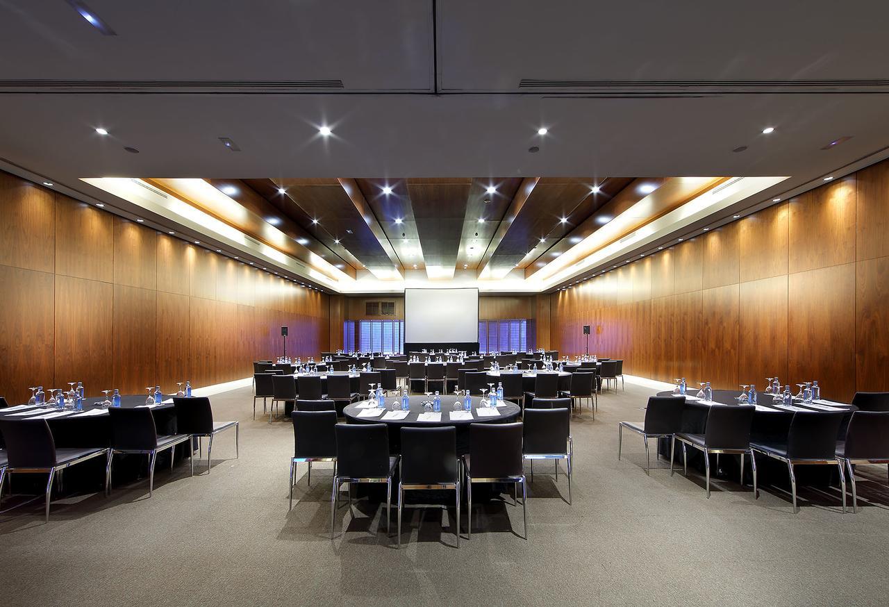 Eurostars Madrid Tower Hotel Exterior photo The photo shows a spacious and modern conference room set up for a meeting or event. There are several round tables arranged in a semi-circle, each covered with a white tablecloth and equipped with water glasses, writing pads, and pens. The room feat
