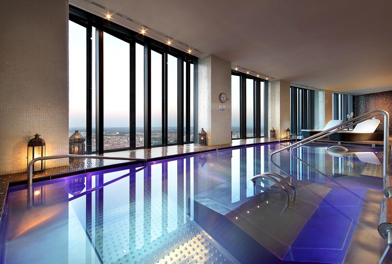 Eurostars Madrid Tower Hotel Exterior photo The photo shows a luxurious indoor pool area with large floor-to-ceiling windows that provide a stunning view of the skyline. The pool features a shiny, reflective surface and has a modern design, with gentle curves in the structure. The lighting is 