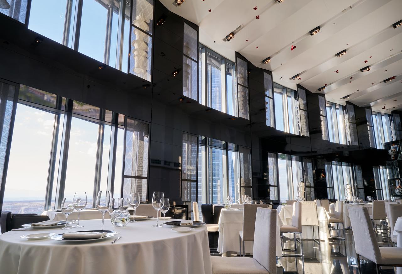 Eurostars Madrid Tower Hotel Exterior photo The photo depicts an elegant restaurant interior characterized by sleek, modern design. It features large floor-to-ceiling windows that allow abundant natural light to fill the space and provide a panoramic view of the outside. The dining area includ