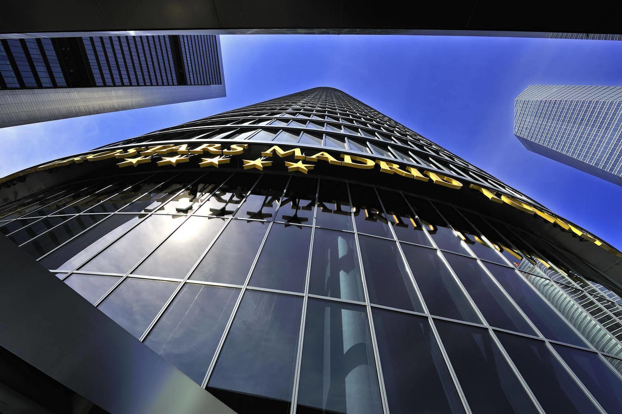 Eurostars Madrid Tower Hotel Exterior photo The photo shows a tall modern building reaching towards the sky. The perspective is from the ground looking upward, highlighting the building's height and glass facade. The building features a large sign that reads "MADRID" in bright yellow letters, 