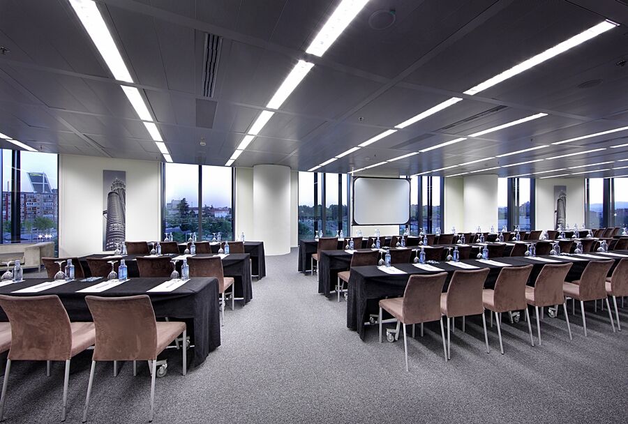 Eurostars Madrid Tower Hotel Exterior photo The photo shows a modern conference room set up for a meeting or seminar. There are multiple rows of tables arranged in a classroom style, each covered with black tablecloths. On the tables, there are notepads and water bottles, suggesting that atten